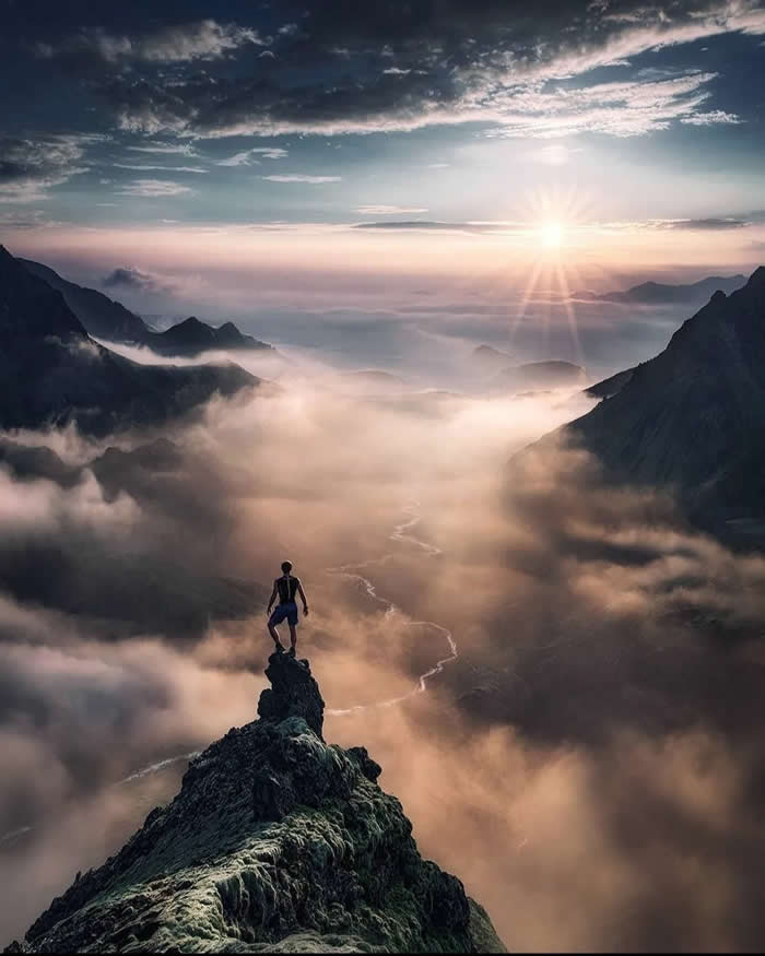 Beauty of the Earth: 33 Epic Landscape Views That Prove Nature Is the Greatest Artist 43 A person stands on a narrow rocky peak above a cloud-filled mountain valley at sunrise, with soft golden light, distant ridges, and a winding river visible through the mist.