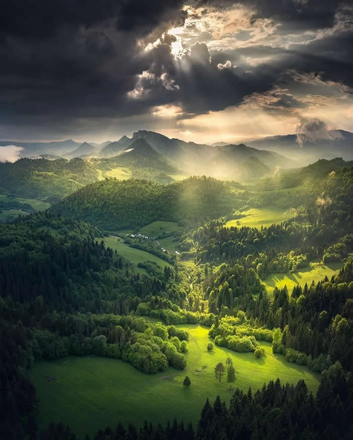 Beauty of the Earth: 33 Epic Landscape Views That Prove Nature Is the Greatest Artist 48 Sun rays stream through dark storm clouds onto a lush green valley filled with forests, rolling hills, and distant mountain ridges, creating bright patches of light across the landscape.