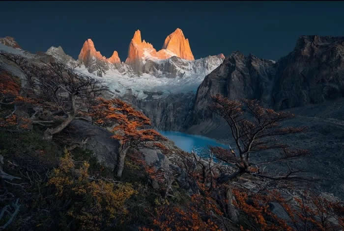 Beauty of the Earth: 33 Epic Landscape Views That Prove Nature Is the Greatest Artist 49 Snow-capped mountain peaks glow orange at sunset above a rocky landscape with twisted trees and a turquoise lake nestled in the valley below.