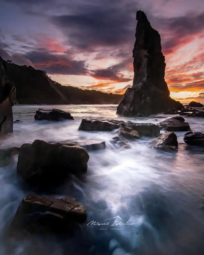 Beauty of the Earth: 33 Epic Landscape Views That Prove Nature Is the Greatest Artist 50 A tall sea stack rises from the ocean at sunset, surrounded by dark rocks and silky long-exposure water beneath a dramatic purple and orange sky.