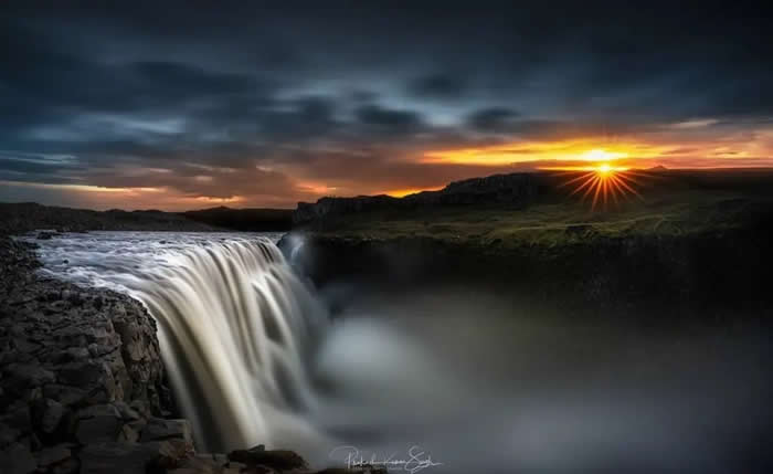 Beauty of the Earth: 33 Epic Landscape Views That Prove Nature Is the Greatest Artist 51 A wide waterfall pours over a rocky cliff at sunset, with silky mist below, dark clouds overhead, and a bright starburst sun glowing on the horizon.