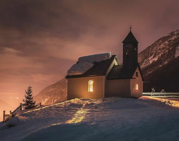 Beauty of the Earth: 33 Epic Landscape Views That Prove Nature Is the Greatest Artist 53 A small chapel with glowing windows stands on a snowy hillside at dusk, with warm light in the sky and dark mountain slopes rising behind it.