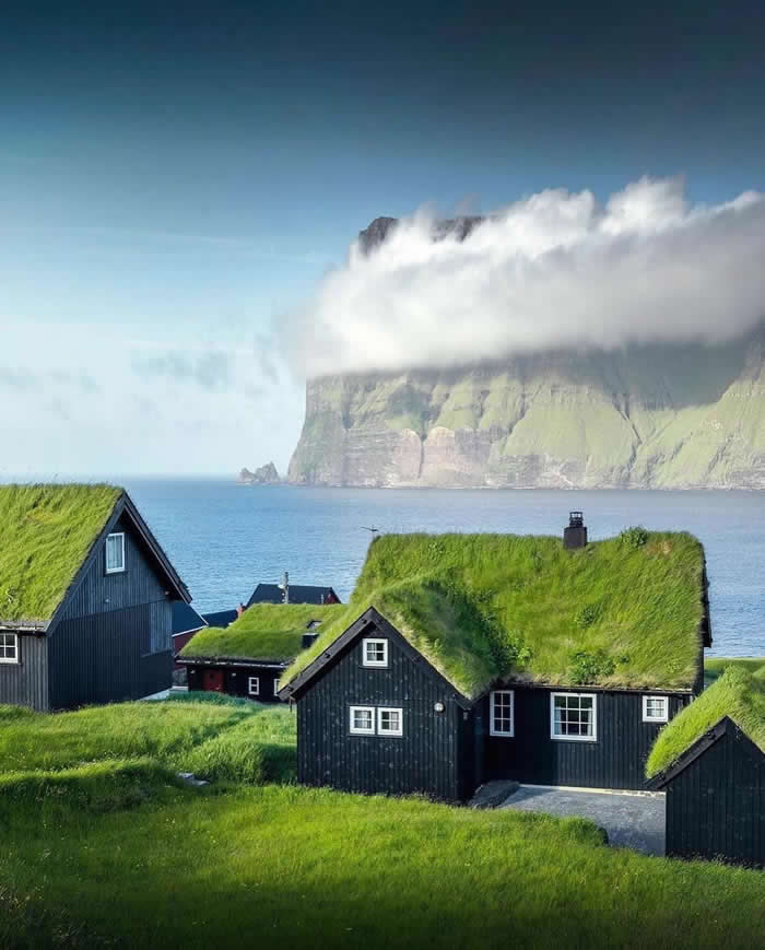 Beauty of the Earth: 33 Epic Landscape Views That Prove Nature Is the Greatest Artist 57 Black wooden houses with green grass roofs sit beside the sea, with a massive cliff partly covered by low clouds rising in the background under a clear blue sky.