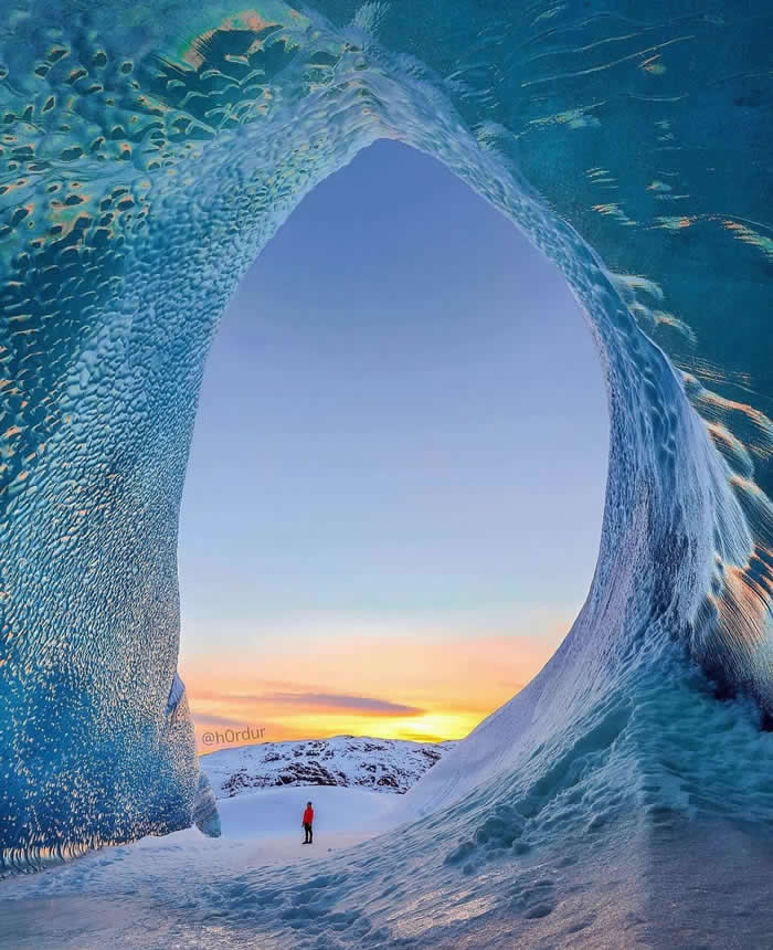 Beauty of the Earth: 33 Epic Landscape Views That Prove Nature Is the Greatest Artist 59 A small person stands on snowy ground beneath a massive blue ice arch, with the opening framing a glowing sunset sky and snow-covered landscape in the distance.