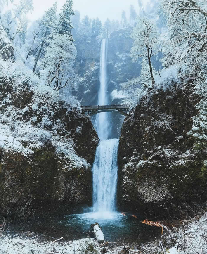 Beauty of the Earth: 33 Epic Landscape Views That Prove Nature Is the Greatest Artist 60 A tall waterfall plunges through a snowy forested gorge, passing beneath a stone bridge before dropping into a pool, with frost-covered trees and dark cliffs surrounding the scene.