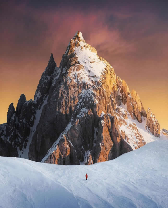 Beauty of the Earth: 33 Epic Landscape Views That Prove Nature Is the Greatest Artist 61 A tiny person in a red jacket stands on a snowy slope beneath a towering rocky mountain peak lit by warm sunset light, with a glowing pink-orange sky in the background.
