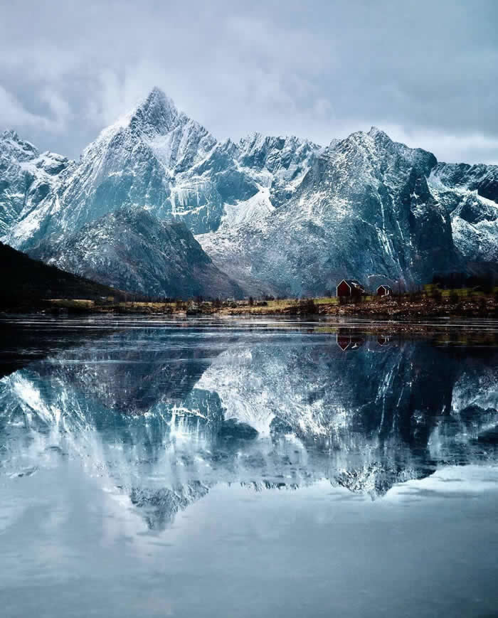 Beauty of the Earth: 33 Epic Landscape Views That Prove Nature Is the Greatest Artist 62 Snow-covered mountains rise above a calm fjord, reflected almost perfectly in the water, with a few small red cabins along the shoreline beneath the towering peaks.