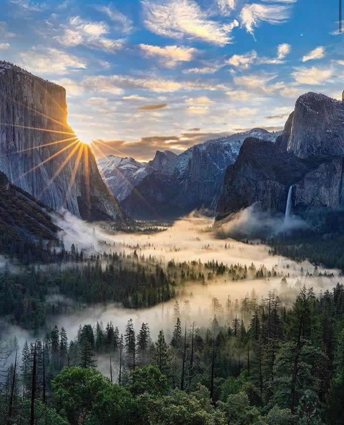 Beauty of the Earth: 33 Epic Landscape Views That Prove Nature Is the Greatest Artist 65 Sunrise lights Yosemite Valley with a starburst beside a towering granite cliff, while mist drifts above the forest below and a waterfall cascades down the opposite rock face.