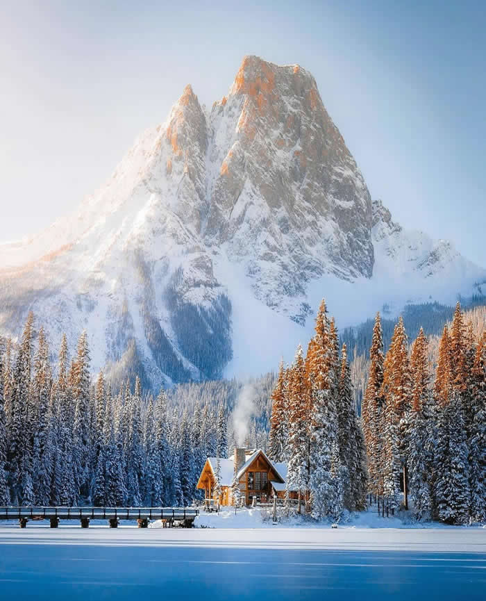 Beauty of the Earth: 33 Epic Landscape Views That Prove Nature Is the Greatest Artist 66 A small wooden cabin sits beside a frozen blue lake and snow-covered pine forest, with a massive sunlit mountain rising dramatically behind under a clear winter sky.