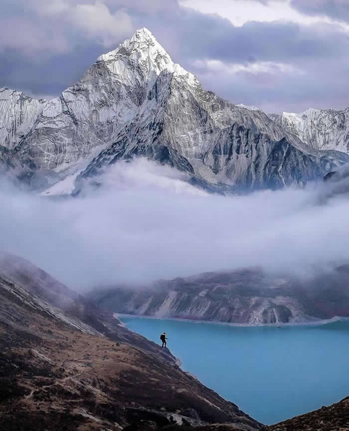 Beauty of the Earth: 33 Epic Landscape Views That Prove Nature Is the Greatest Artist 67 A lone hiker stands on a rocky slope above a bright turquoise lake, with low clouds drifting beneath a towering snow-covered mountain under a soft blue and lavender sky.