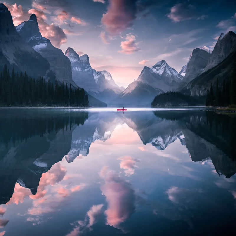 A lone red canoe floats on a perfectly still lake in the Canadian Rockies at sunrise, with snow-capped mountains and pink clouds reflected flawlessly in the calm water.