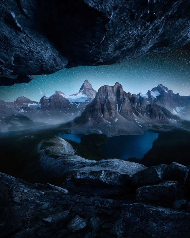 A star-filled night sky illuminates the Canadian Rockies, viewed through the opening of a rocky cave overlooking a deep blue alpine lake and dramatic snow-capped mountain peaks.