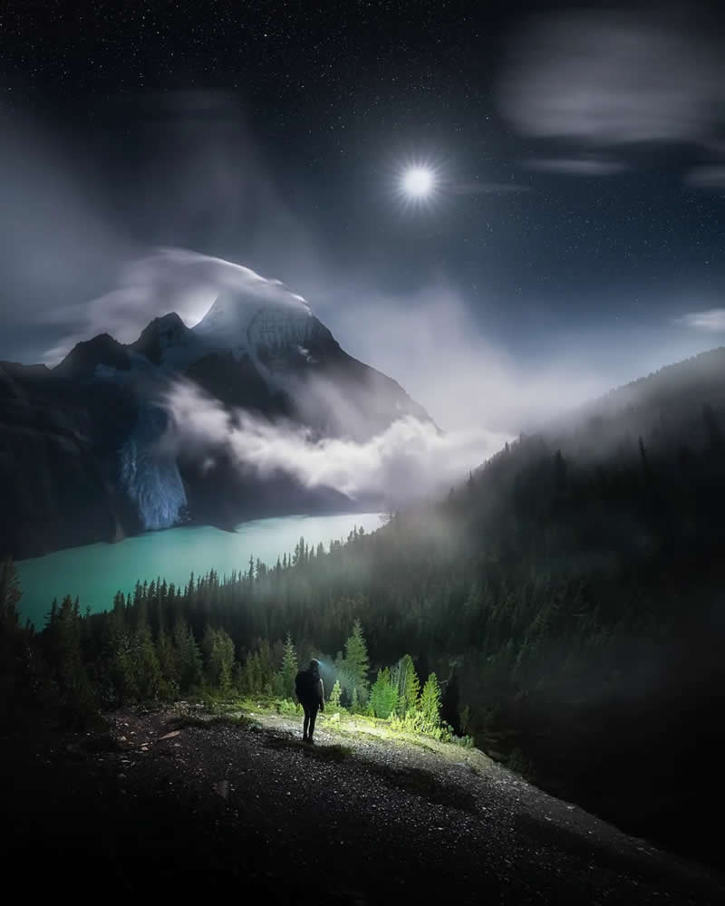 A lone hiker overlooks a turquoise glacial lake beneath moonlit, mist-covered mountains in the Canadian Rockies, with evergreen forests and a star-filled night sky creating a dramatic alpine landscape.