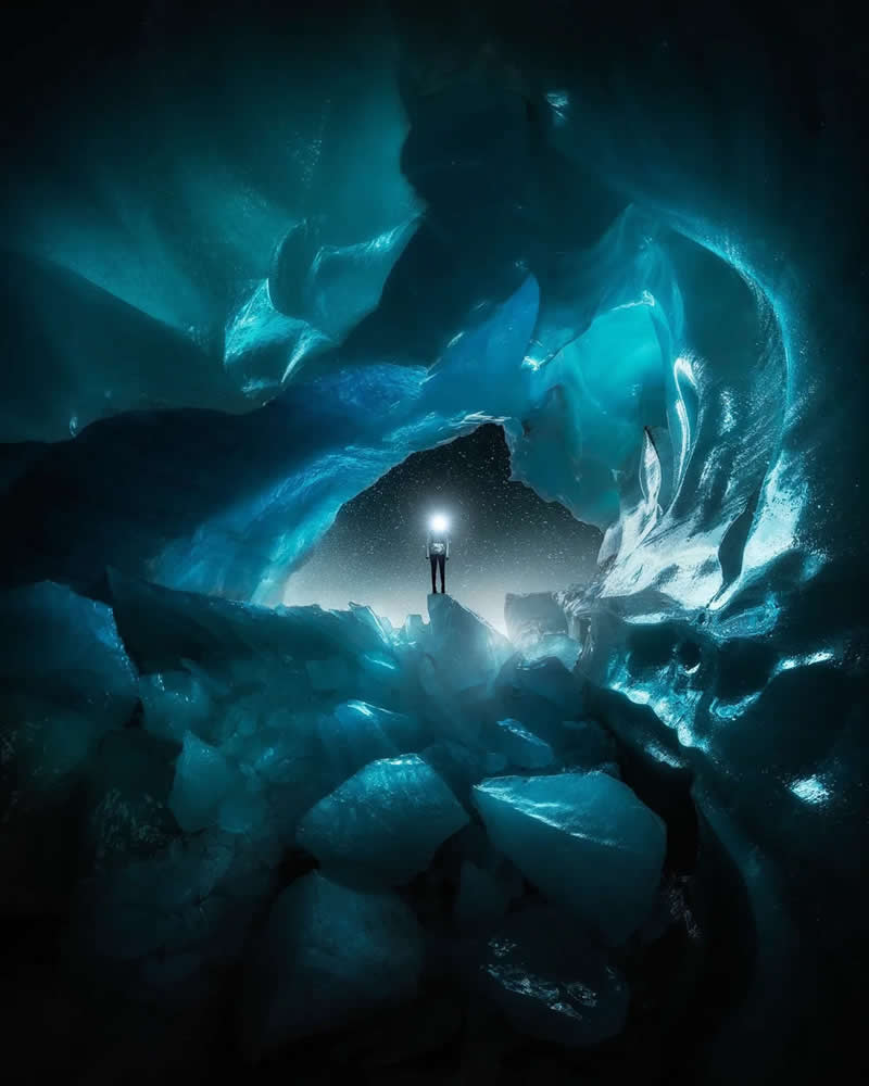 A solitary explorer with a bright headlamp stands inside a glowing blue ice cave, surrounded by massive sculpted ice formations and crystalline textures in a dramatic frozen landscape.