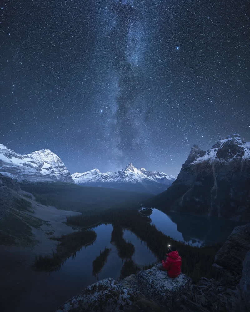 A lone person in a red jacket sits on a rocky overlook beneath the Milky Way, gazing across winding lakes and snow-capped peaks in the Canadian Rockies at night.