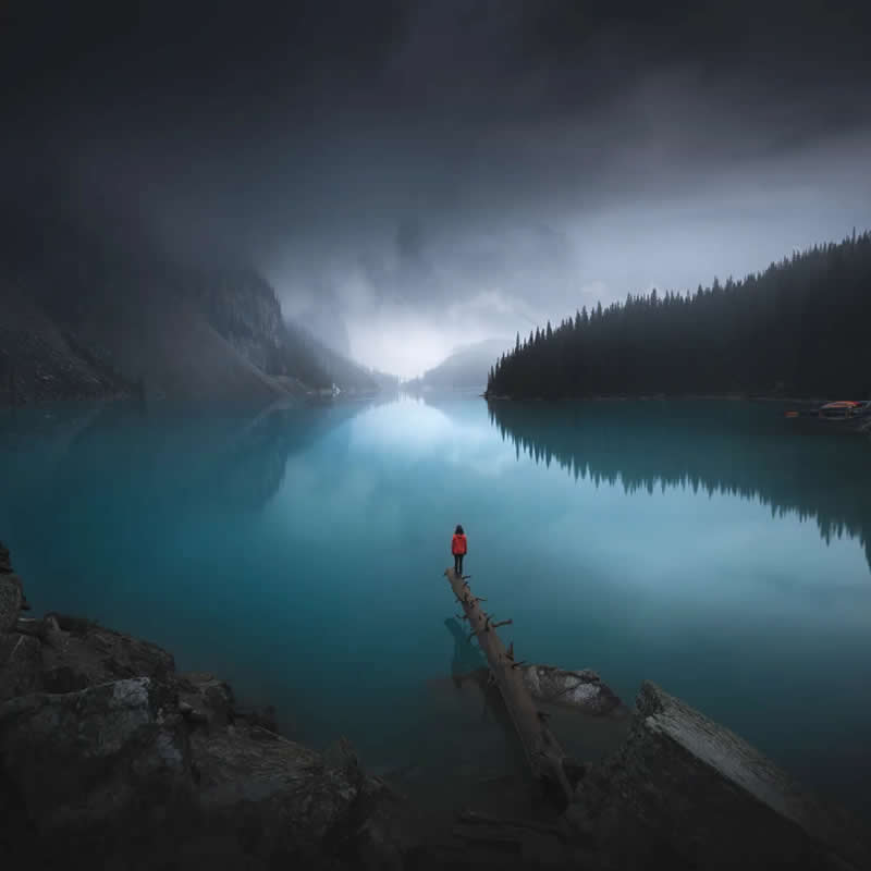 A lone person in a red jacket stands on a fallen log extending into a turquoise glacial lake, surrounded by misty mountains, evergreen forests, and calm reflections in the Canadian Rockies.