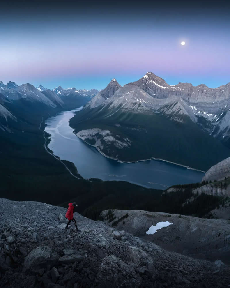 A lone hiker in a red jacket ascends a rocky slope overlooking a winding alpine lake, surrounded by towering Canadian Rockies beneath a pastel twilight sky and a glowing moon.