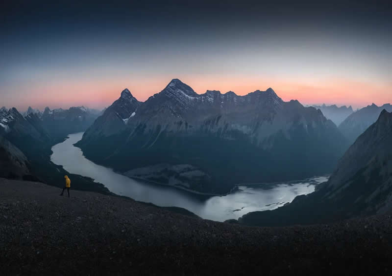 A lone hiker in a yellow jacket walks across a rocky ridge overlooking winding lakes and towering mountain peaks at dawn in the Canadian Rockies beneath a soft pastel sky.
