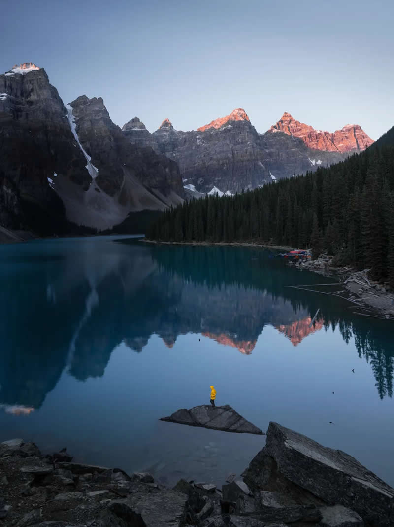 A person in a yellow jacket stands on a rock in a calm turquoise lake, reflecting sunlit mountain peaks, evergreen forests, and the serene beauty of the Canadian Rockies at sunrise.