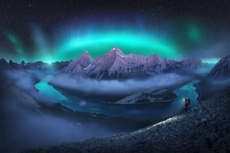 A lone hiker stands on a rocky ridge beneath the northern lights, overlooking winding rivers, mist-covered valleys, and dramatic mountain peaks in the Canadian Rockies under a star-filled night sky.