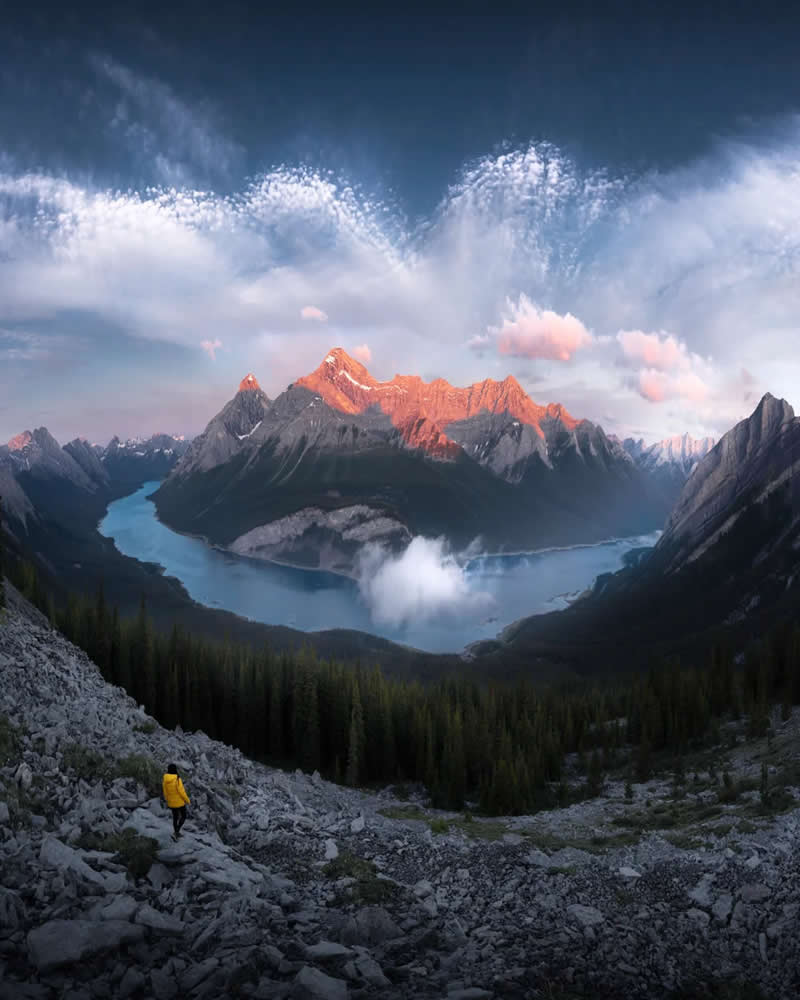 A lone hiker in a yellow jacket walks down a rocky mountainside overlooking winding turquoise lakes and sunrise-lit peaks beneath dramatic clouds in the Canadian Rockies.