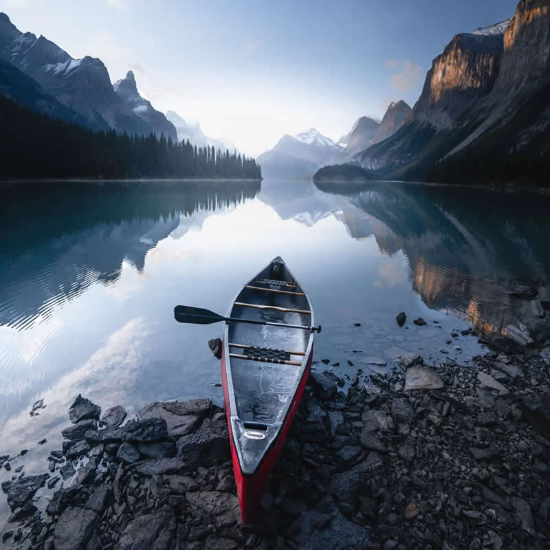 A red canoe rests on the rocky shore of a calm alpine lake, reflecting towering mountains and evergreen forests beneath a clear morning sky in the Canadian Rockies.