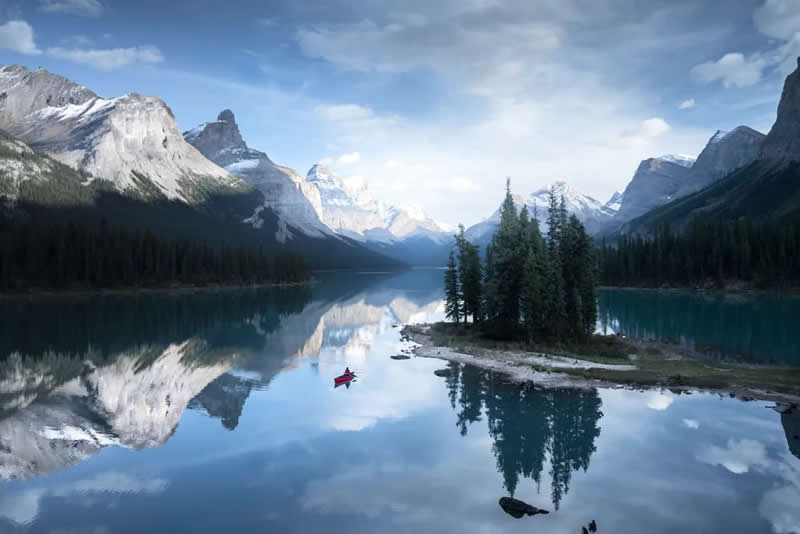 A lone red canoe floats beside a small tree-covered island on a calm alpine lake, reflecting snow-capped mountains, evergreen forests, and a peaceful sky in the Canadian Rockies.