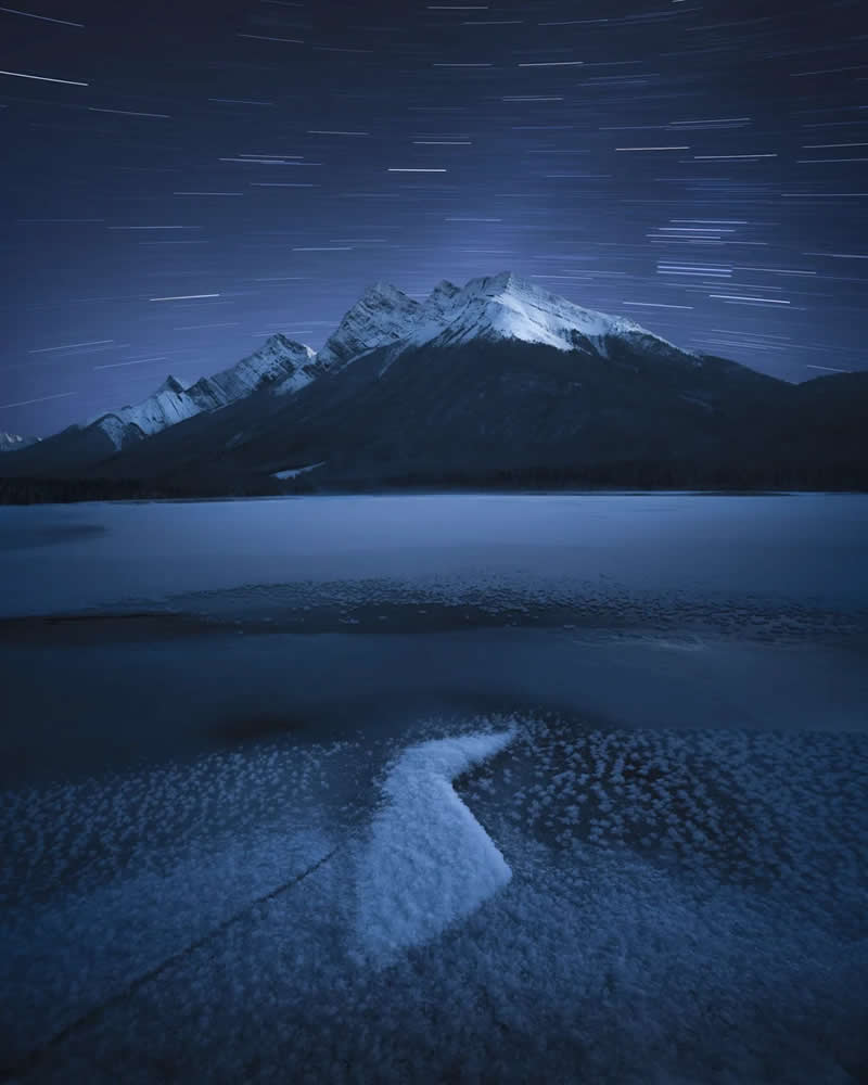 Star trails arc across the night sky above snow-covered mountain peaks and a frozen alpine lake, with textured ice in the foreground creating a serene Canadian Rockies winter landscape.