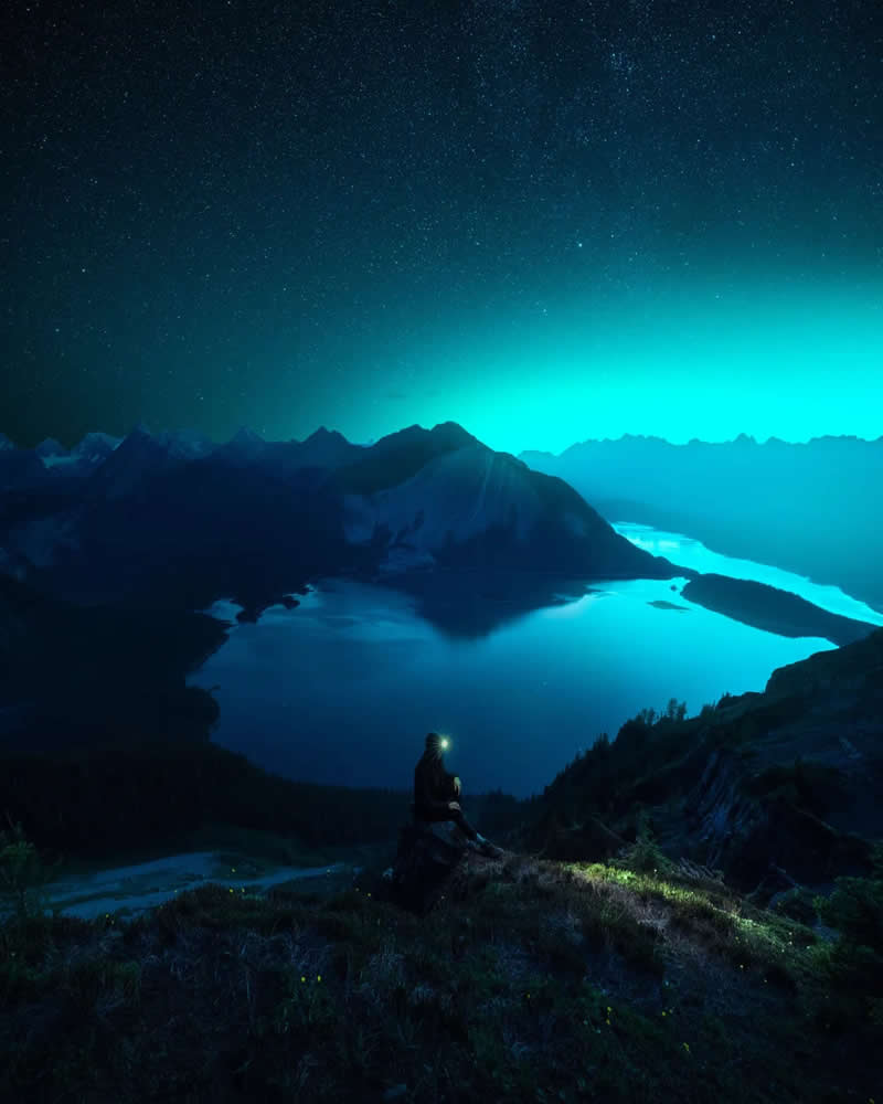 A lone hiker with a headlamp overlooks glowing alpine lakes and dark mountain silhouettes beneath a star-filled night sky, creating a peaceful and atmospheric Canadian Rockies landscape.