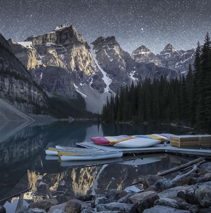 Colorful canoes rest on a wooden dock beside a calm alpine lake, reflecting towering snow-capped mountains, evergreen forests, and a star-filled night sky in the Canadian Rockies.