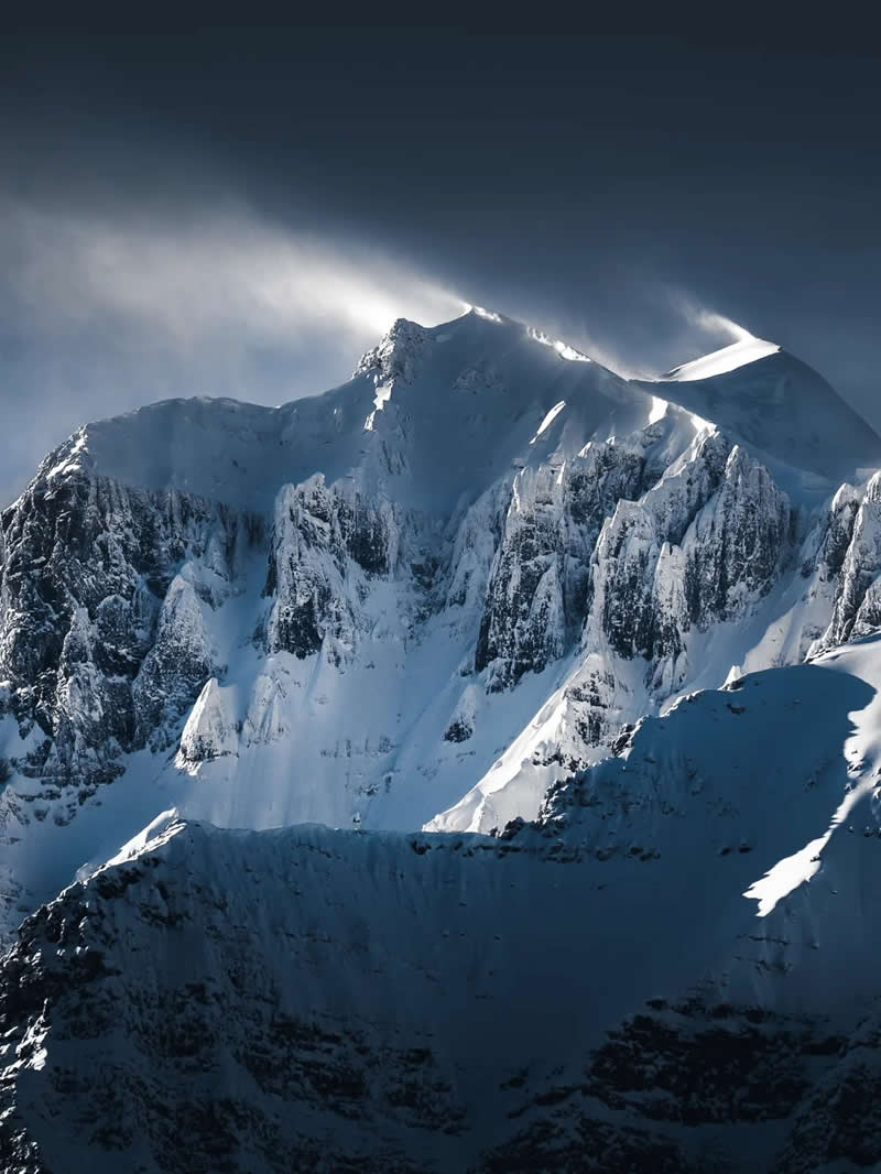A close-up view of snow-covered mountain peaks with windblown spindrift sweeping across the summits, highlighting rugged cliffs, icy textures, and dramatic light in the Canadian Rockies.
