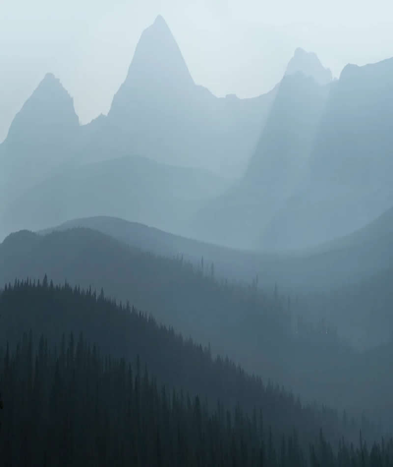 Misty mountain peaks rise above layered forested ridges in the Canadian Rockies, with soft atmospheric haze creating depth, silhouettes, and a peaceful minimalist landscape.