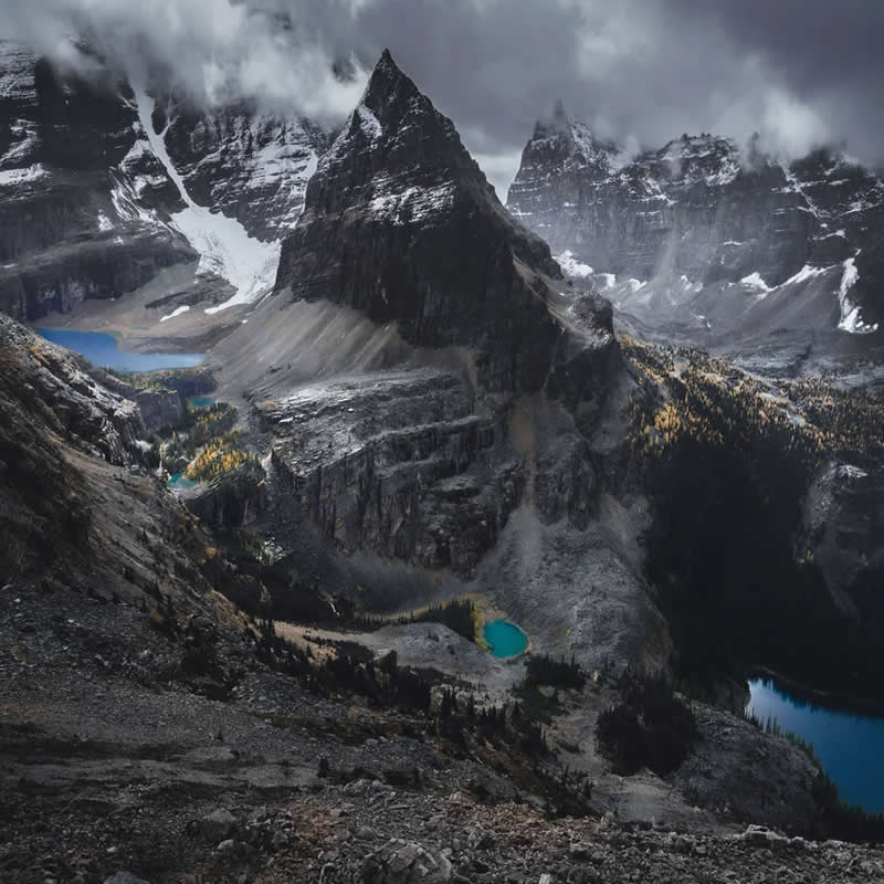 Towering jagged mountain peaks rise above turquoise glacial lakes and rocky valleys, with low clouds swirling around snow-covered summits in the dramatic Canadian Rockies landscape.