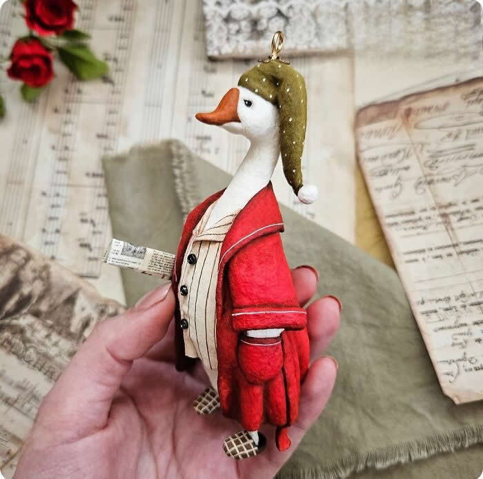 Cotton Wool Sculptures. A handcrafted miniature goose ornament made from cotton wool is dressed in a deep red coat and striped vest, wearing a soft green nightcap while holding a tiny rolled paper scroll against a vintage-style holiday background.
