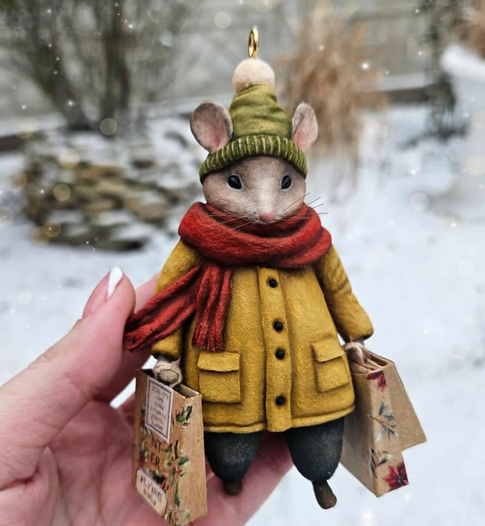 A handcrafted miniature mouse ornament made from cotton wool wears a mustard-yellow coat, green knit hat, and deep red scarf while holding tiny shopping bags against a softly blurred snowy background.