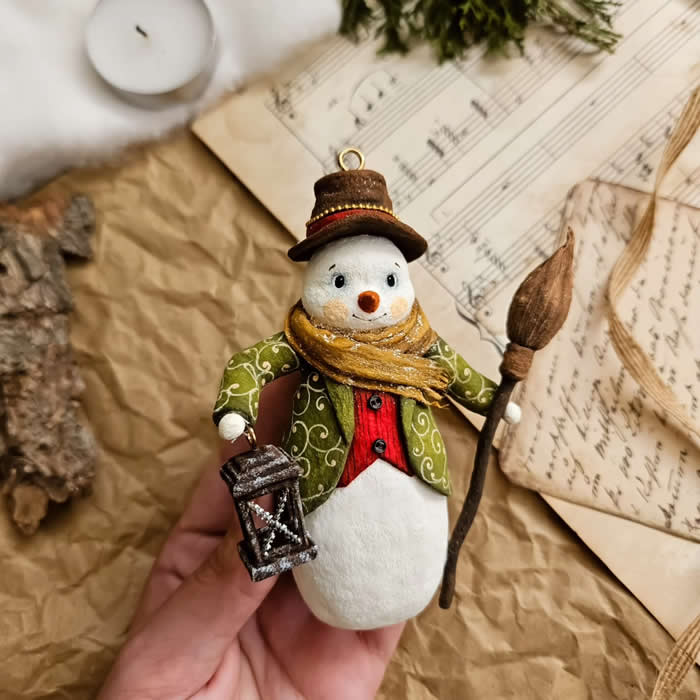 Cotton Wool Sculptures. A handcrafted miniature snowman ornament made from cotton wool wears a green patterned coat, red vest, brown hat, and golden scarf while holding a lantern and a rustic broom against a vintage paper and sheet music backdrop.