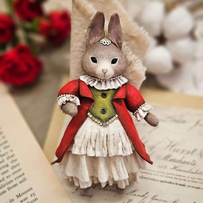 A handcrafted miniature rabbit ornament made from cotton wool wears a rich red coat, olive-green bodice, and layered cream dress with delicate ruffled details, posed against a soft vintage-inspired background.