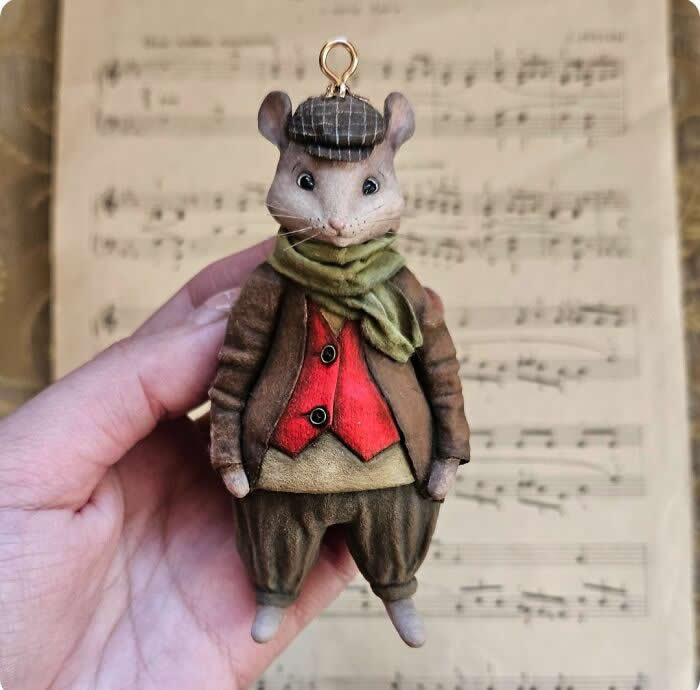 A handcrafted miniature mouse ornament made from cotton wool wears a brown coat, red waistcoat, olive scarf, and plaid cap, standing against a softly blurred sheet music background with a charming old-world feel.