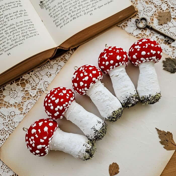 Five handcrafted fly agaric mushroom ornaments made from cotton wool are arranged in a row on a lace-covered surface, with bright red caps, white speckles, and textured stems styled beside an open vintage book.