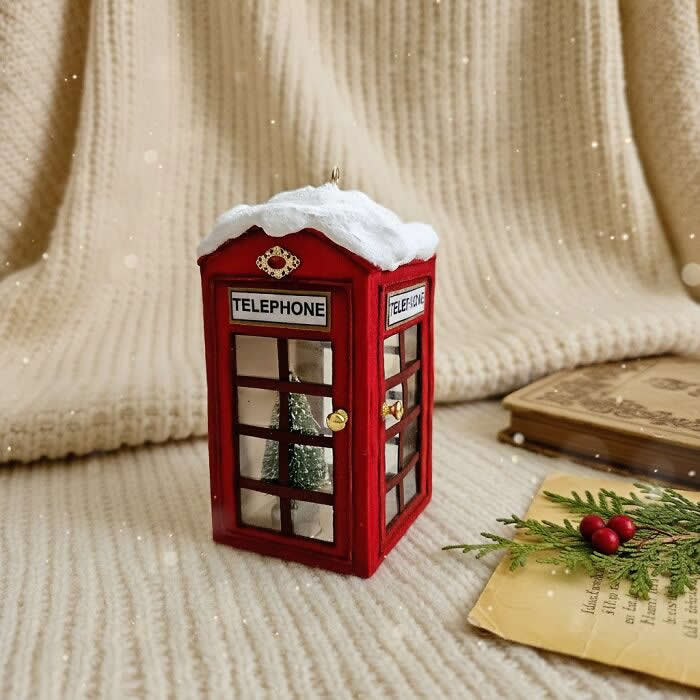 A handcrafted miniature red telephone booth ornament made from cotton wool stands on a cozy knitted backdrop, topped with snow and featuring tiny window details with a small tree visible inside.