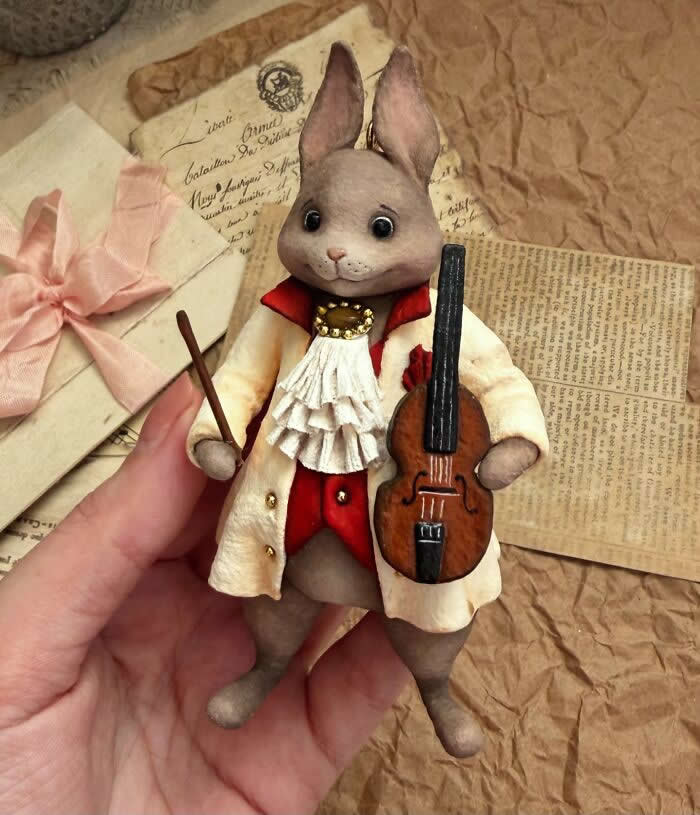 A handcrafted miniature rabbit ornament made from cotton wool wears an elegant cream coat with red accents and a ruffled cravat, holding a tiny violin and bow against a vintage paper background.