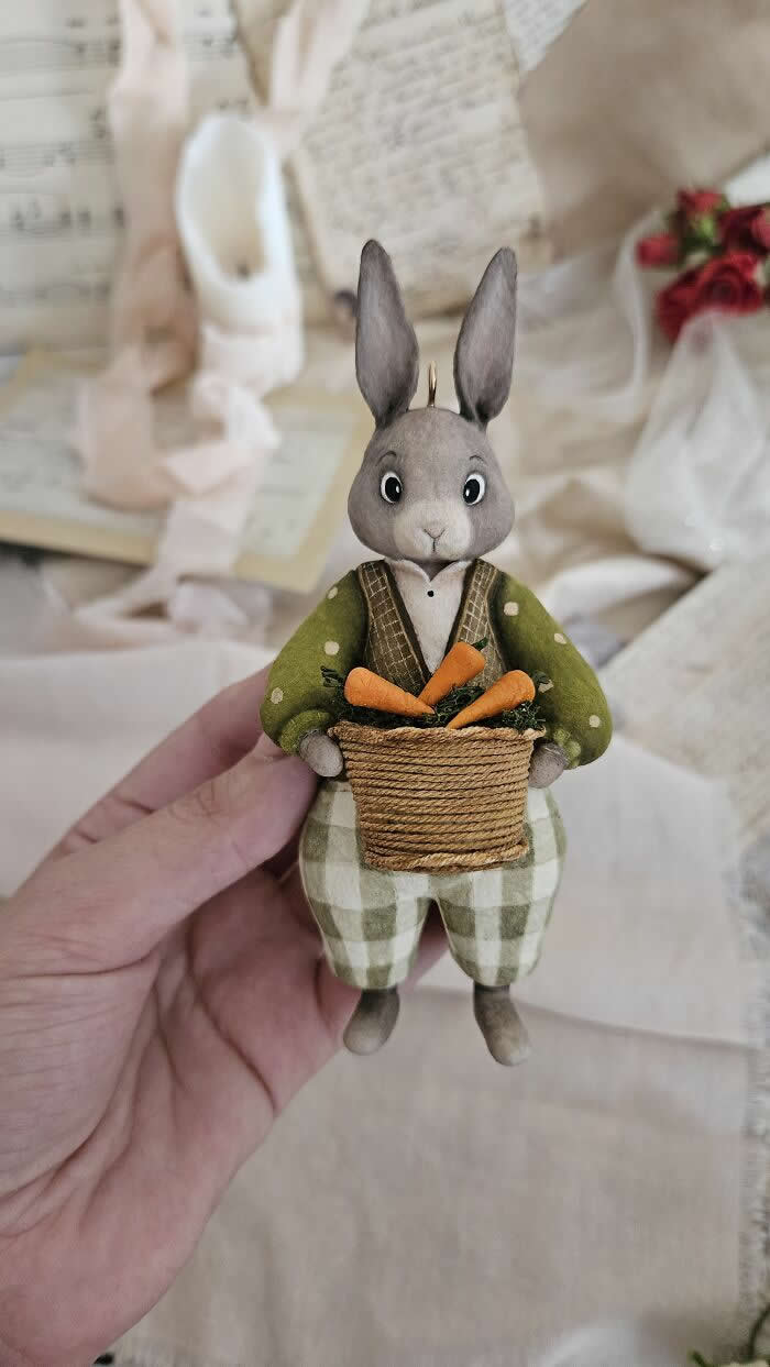 A handcrafted miniature rabbit ornament made from cotton wool wears a soft green jacket and checkered trousers while holding a woven basket filled with tiny carrots, posed against a delicate vintage-inspired background.