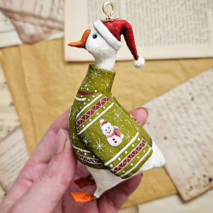 A handcrafted miniature goose ornament made from cotton wool wears a cozy olive-green Christmas sweater with festive patterns and a tiny snowman design, paired with a red Santa hat, posed against a vintage paper background.