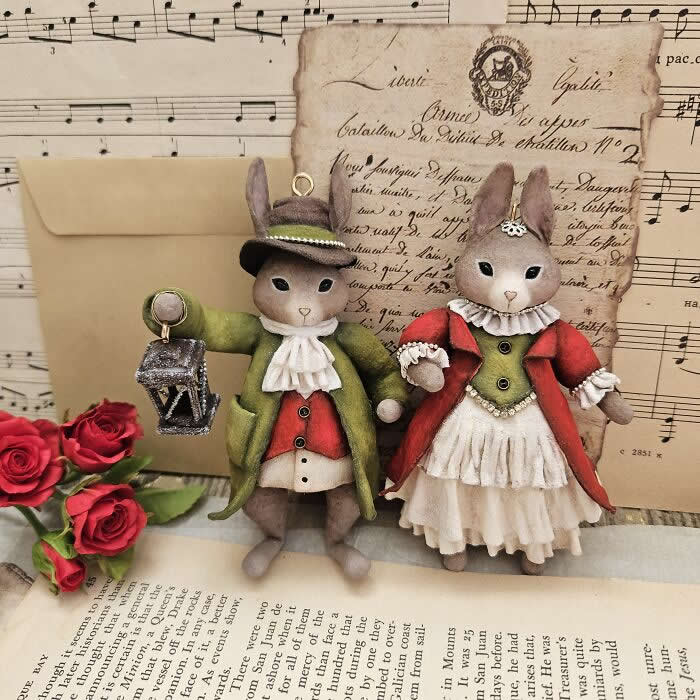 Two handcrafted miniature rabbit ornaments made from cotton wool stand together against a vintage backdrop of sheet music, old letters, and red roses&mdash;one dressed in a green coat holding a lantern, the other in a red coat and layered cream dress.