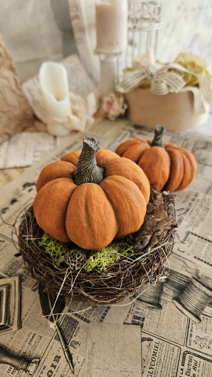 Handcrafted miniature pumpkin ornaments made from cotton wool sit in a rustic twig nest with moss, styled on vintage newspaper pages beside candles and soft autumn d&eacute;cor in a warm, nostalgic setting.
