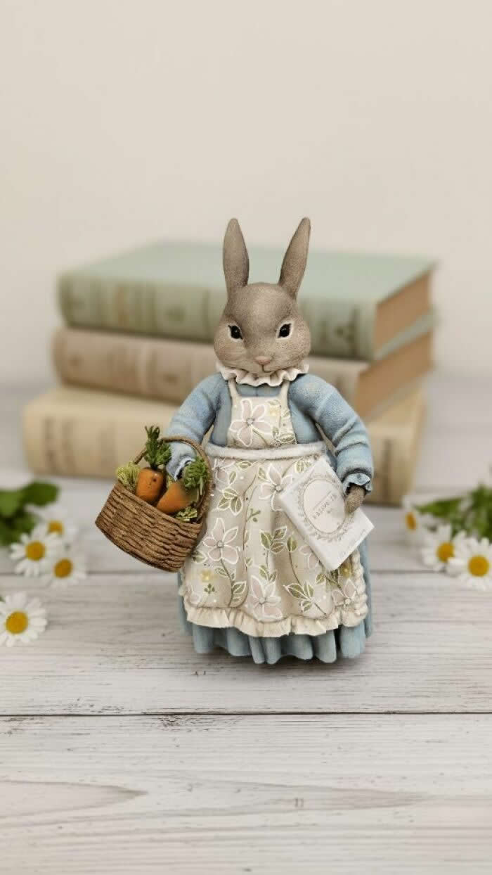 A handcrafted miniature rabbit ornament made from cotton wool wears a soft blue dress and floral apron, carrying a woven basket of tiny carrots and a small letter, posed in front of stacked vintage books and scattered daisies.