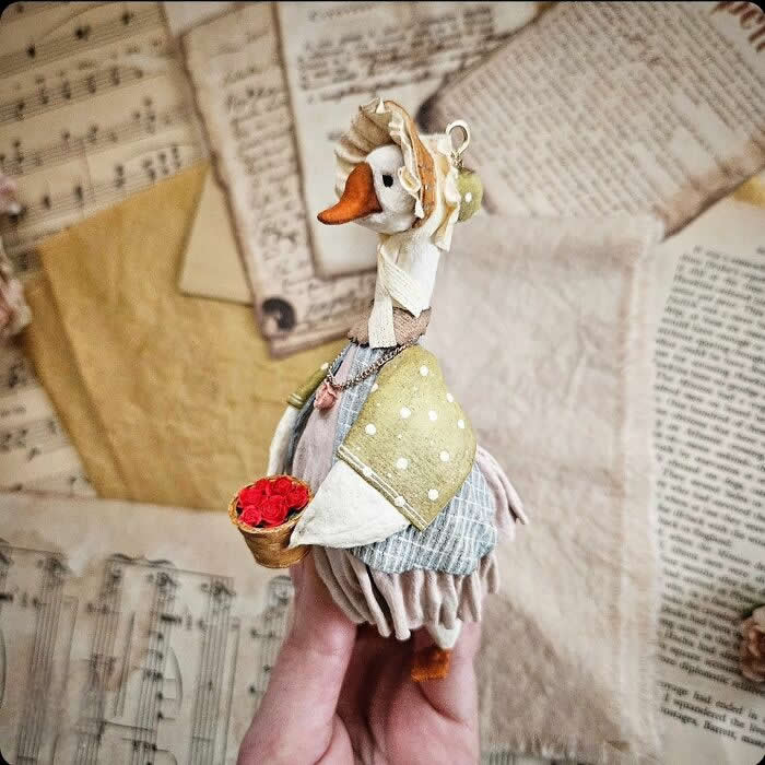 A handcrafted miniature goose ornament made from cotton wool wears a soft bonnet, polka-dot shawl, and layered vintage dress while carrying a tiny basket of red roses, posed against a backdrop of old sheet music and aged paper.