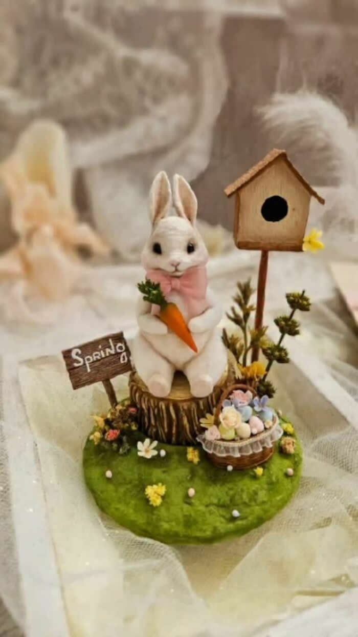 A handcrafted miniature white rabbit ornament made from cotton wool sits on a mossy spring scene holding a carrot, surrounded by tiny flowers, pastel eggs, a birdhouse, and a wooden &ldquo;Spring&rdquo; sign.