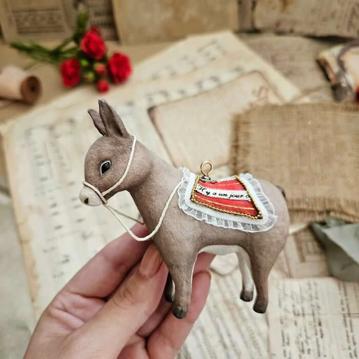 A handcrafted miniature donkey ornament made from cotton wool stands in profile with a delicate bridle and a decorative saddle blanket trimmed with lace, photographed against a vintage backdrop of sheet music, old letters, and red roses.