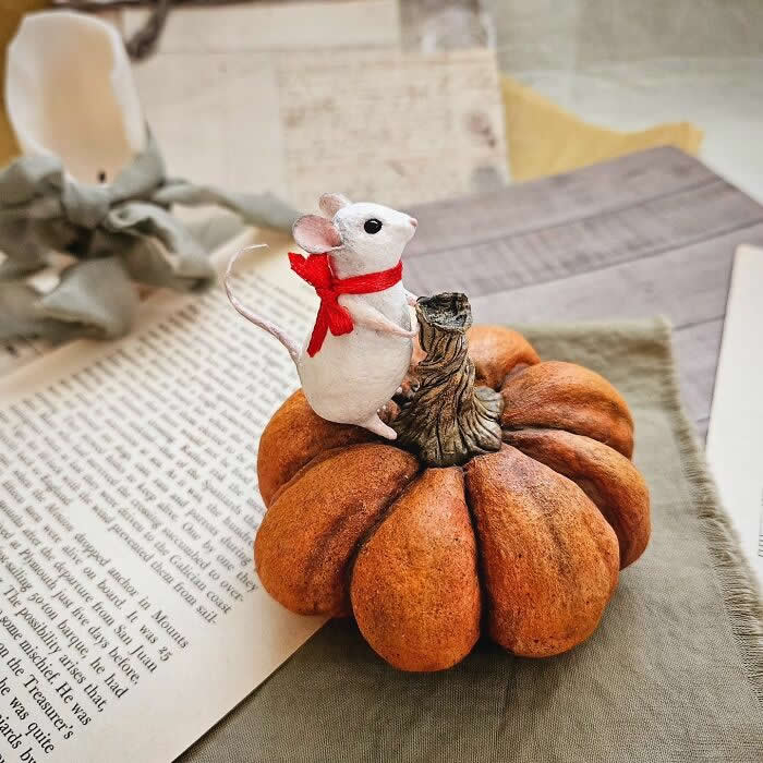 A handcrafted miniature mouse ornament made from cotton wool stands on a rustic pumpkin, wearing a bright red scarf and posed beside vintage book pages and soft autumn d&eacute;cor.